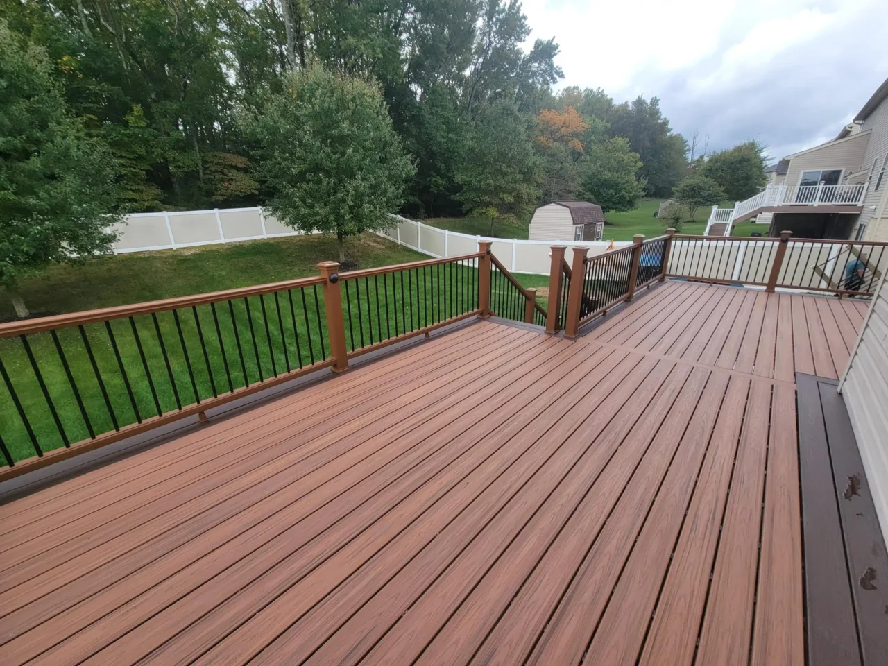 a composite deck with brown flooring and a railing with black balusters and a brown top rail. In the background, a green lawn, trees, and a white fence are visible. A small shed is visible beyond the fence.