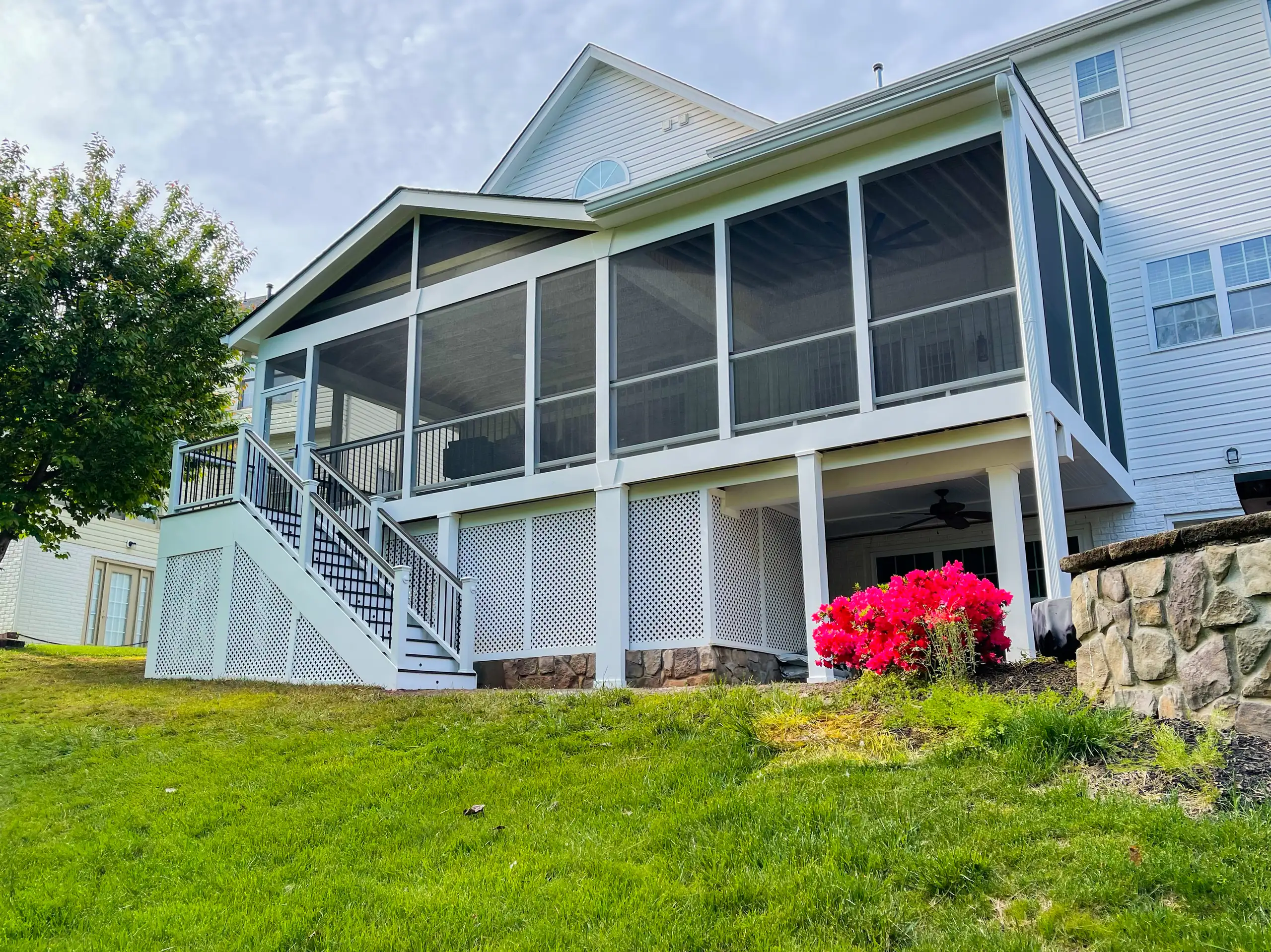 Temporary Patio Enclosures for Winter A large, elevated screened-in porch addition attached to a white suburban home. The structure features a pitched gable roof that integrates seamlessly with the main house, complemented by white railing and staircases, as well as decorative white lattice skirting that conceals the high foundation on the sloping lawn.