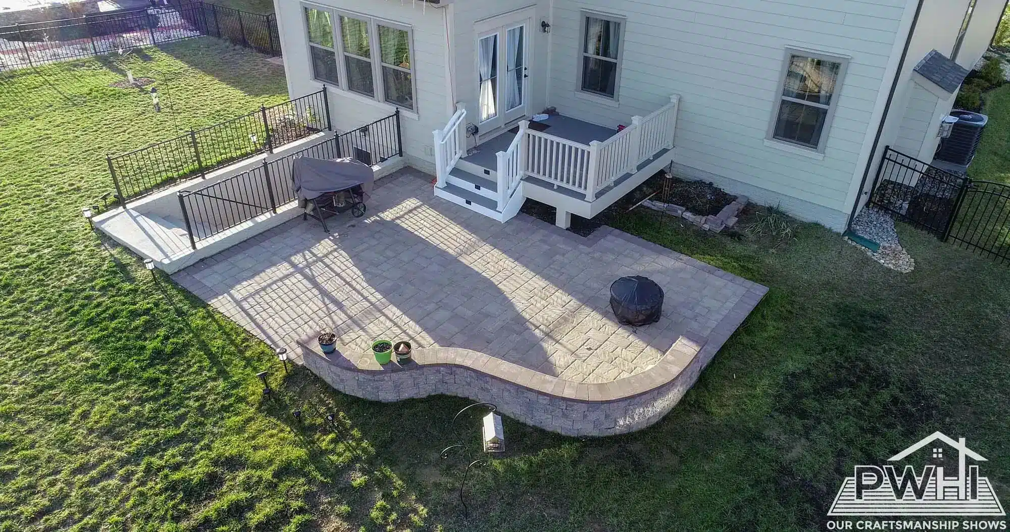 An aerial view shows a newly installed stone paver patio and walkway next to a light-colored house. The patio includes a curved retaining wall and a fire pit, with a short set of white stairs leading down from a back door. A separate concrete walkway leads to a basement entrance.