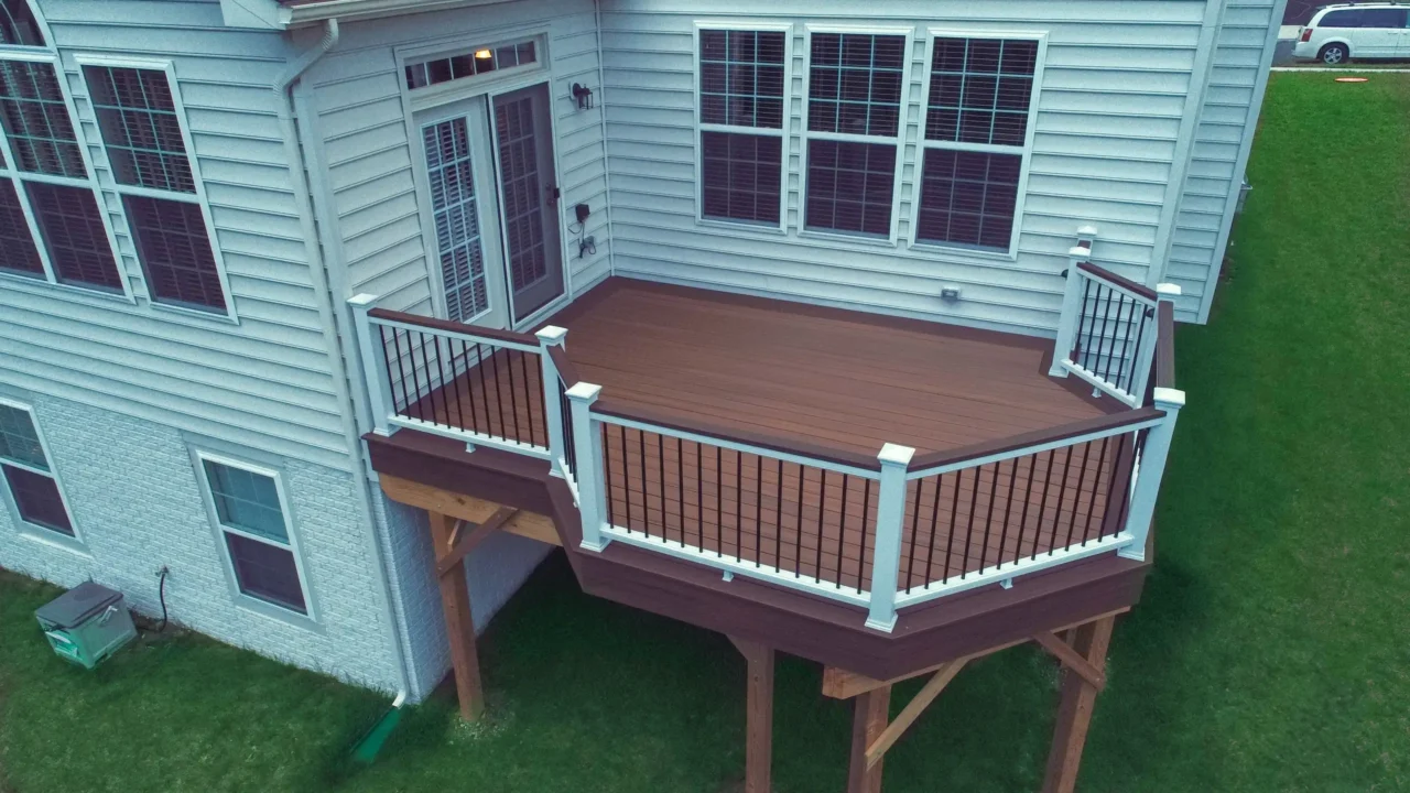 An elevated, angled deck attached to a light gray, vinyl-sided house. The deck features dark brown composite decking and a white railing system with black balusters. A glass French door on the upper level of the house provides access to the deck.