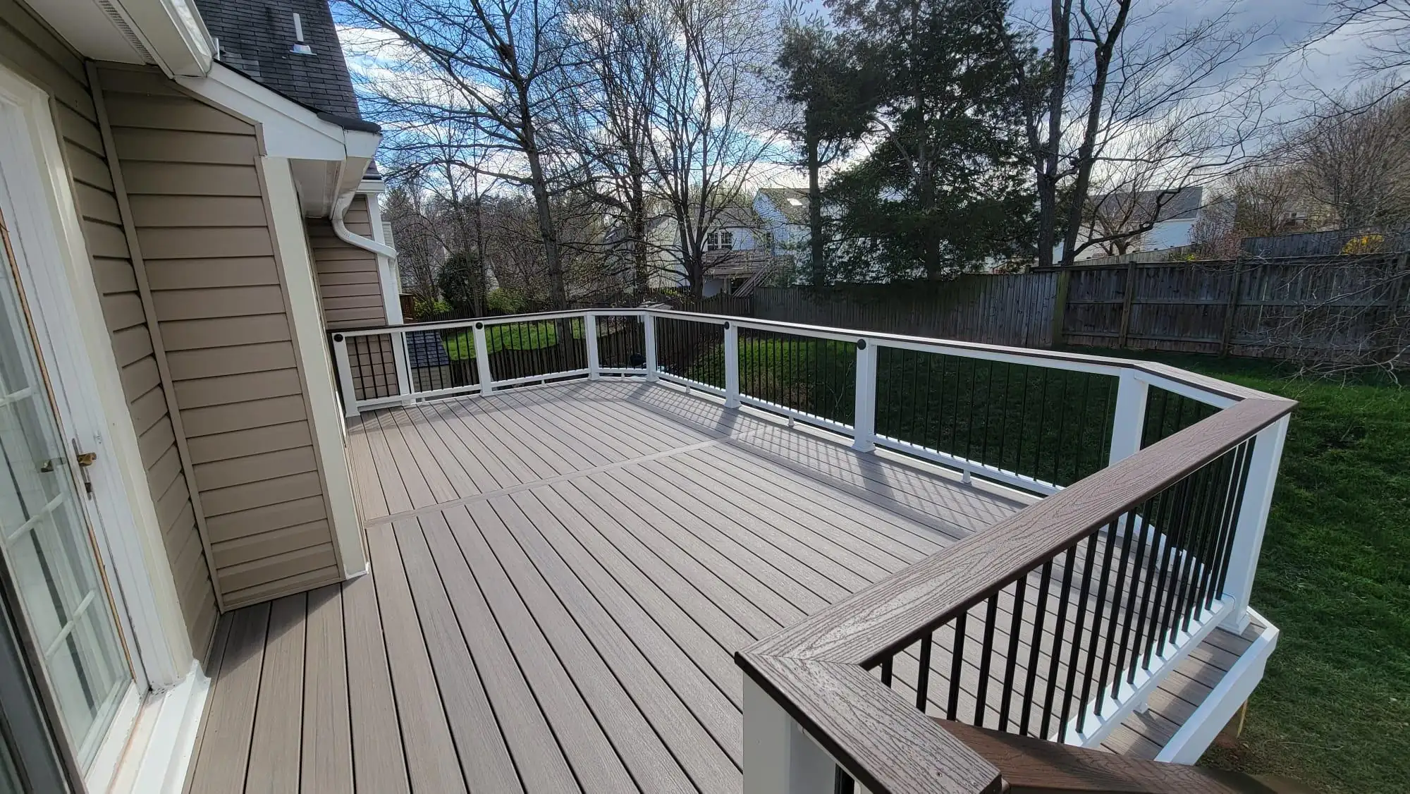composite decking builder in ashburn va A large composite deck with light gray-brown planks extends from a tan-sided house. The deck features clipped corners and is enclosed by a white railing with black metal balusters and a dark brown top rail. A sliding glass door provides access to the house.