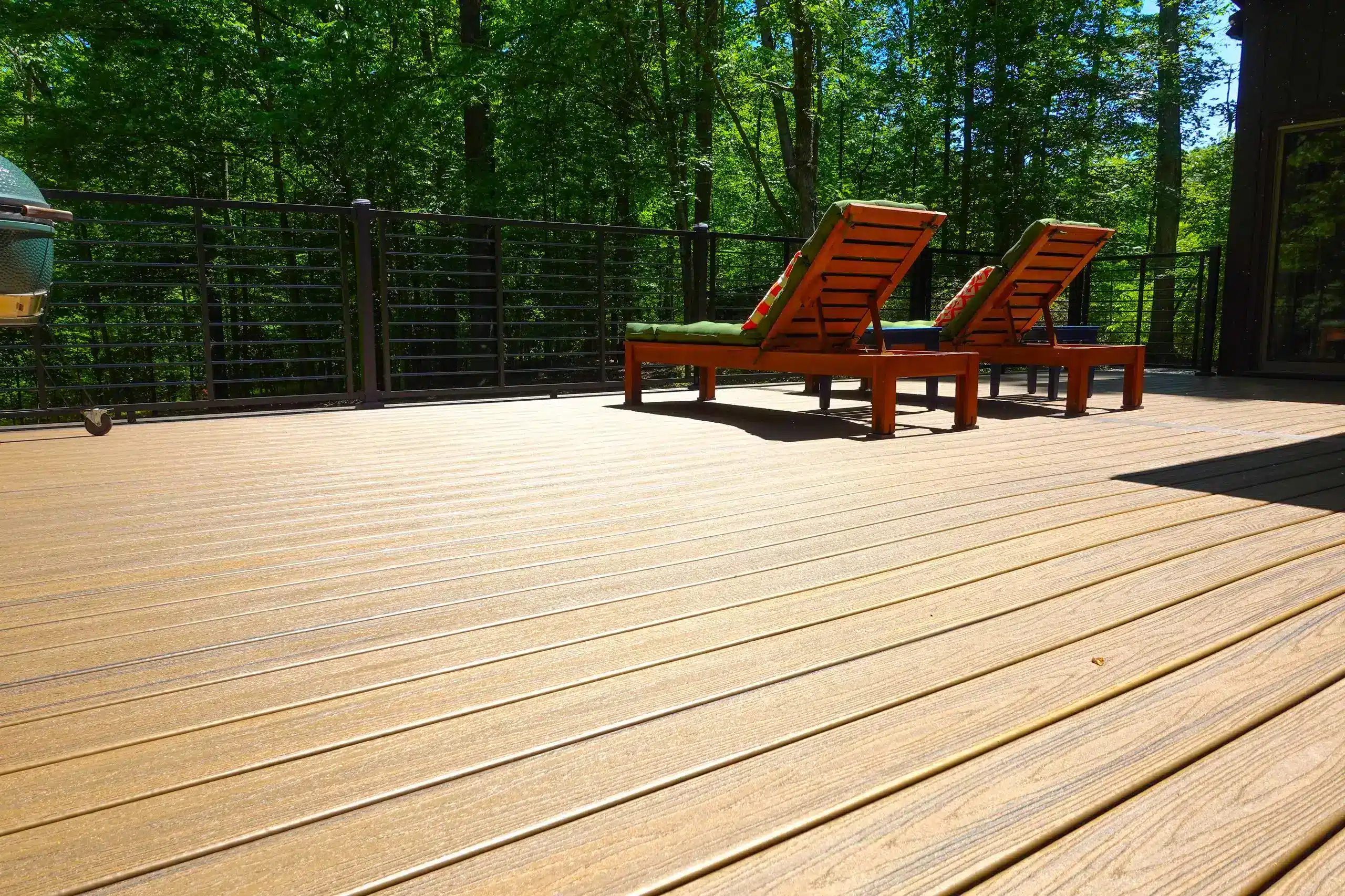A sunny outdoor deck made of light-brown composite planks features two wooden chaise lounges with green-and-orange patterned cushions. A horizontal black metal cable railing encloses the deck and overlooks a lush, dense wooded area.