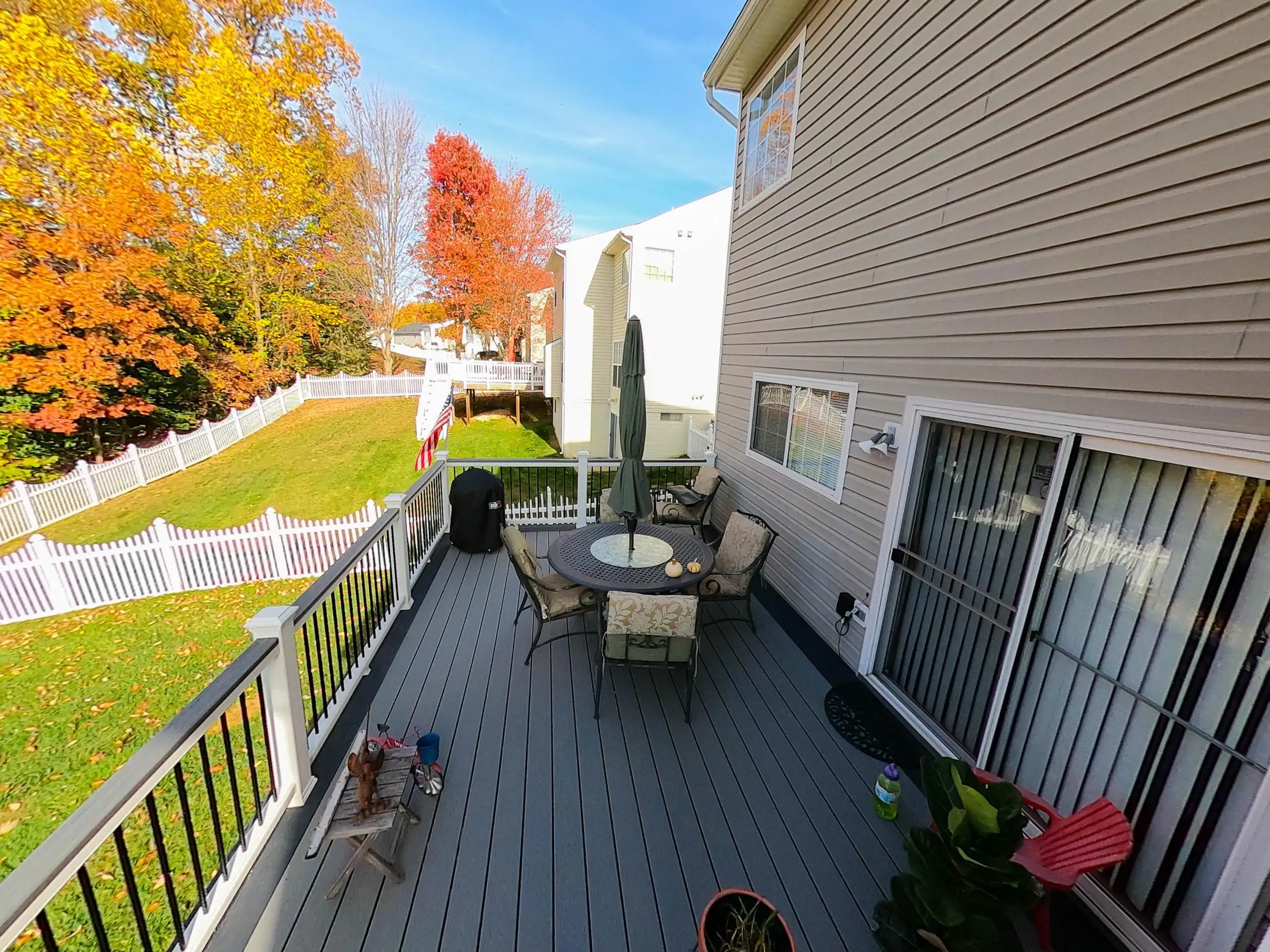 A gray composite deck extends from a tan-sided house on a sunny autumn day. The deck has a white railing with black balusters. Furniture includes a round dining table with an umbrella, four chairs, and a black gas grill. It overlooks a yard with a white picket fence and trees with yellow and red fall foliage.