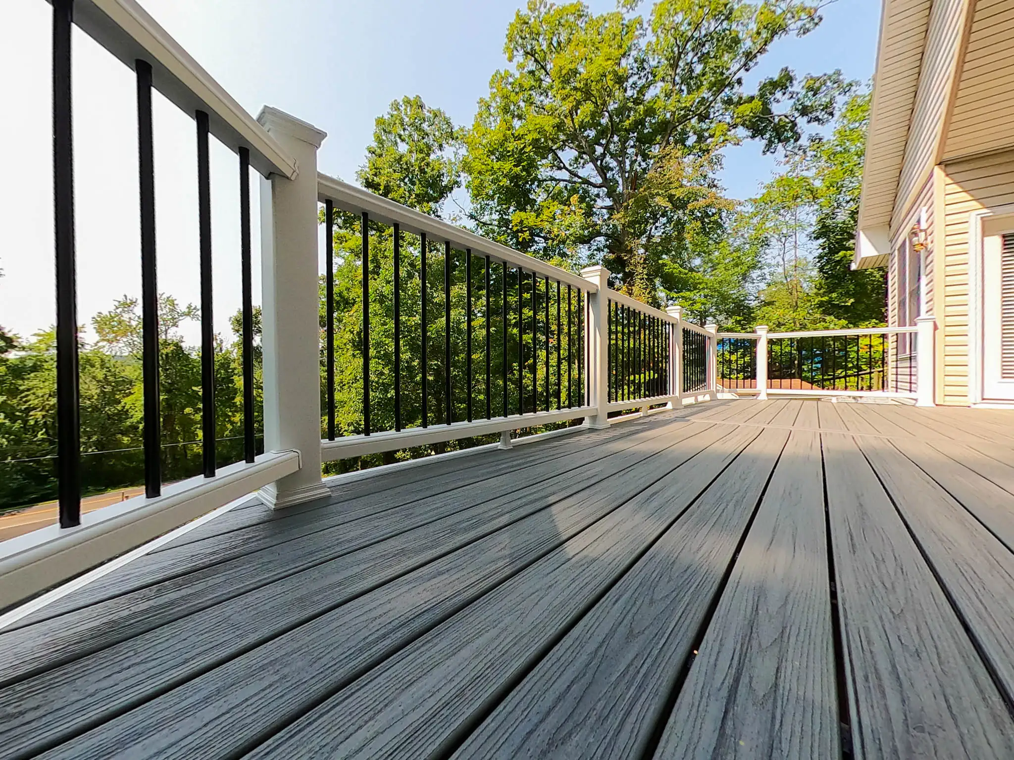 composite decking installation in ashburn va A ground-level view of a wide composite deck with dark gray planks, with the lines of the planks drawing the eye into the distance. The deck is enclosed by a white railing with black vertical balusters and a black top rail. The deck extends along the side of a light-colored house with beige siding, where a white sliding glass door is visible on the right