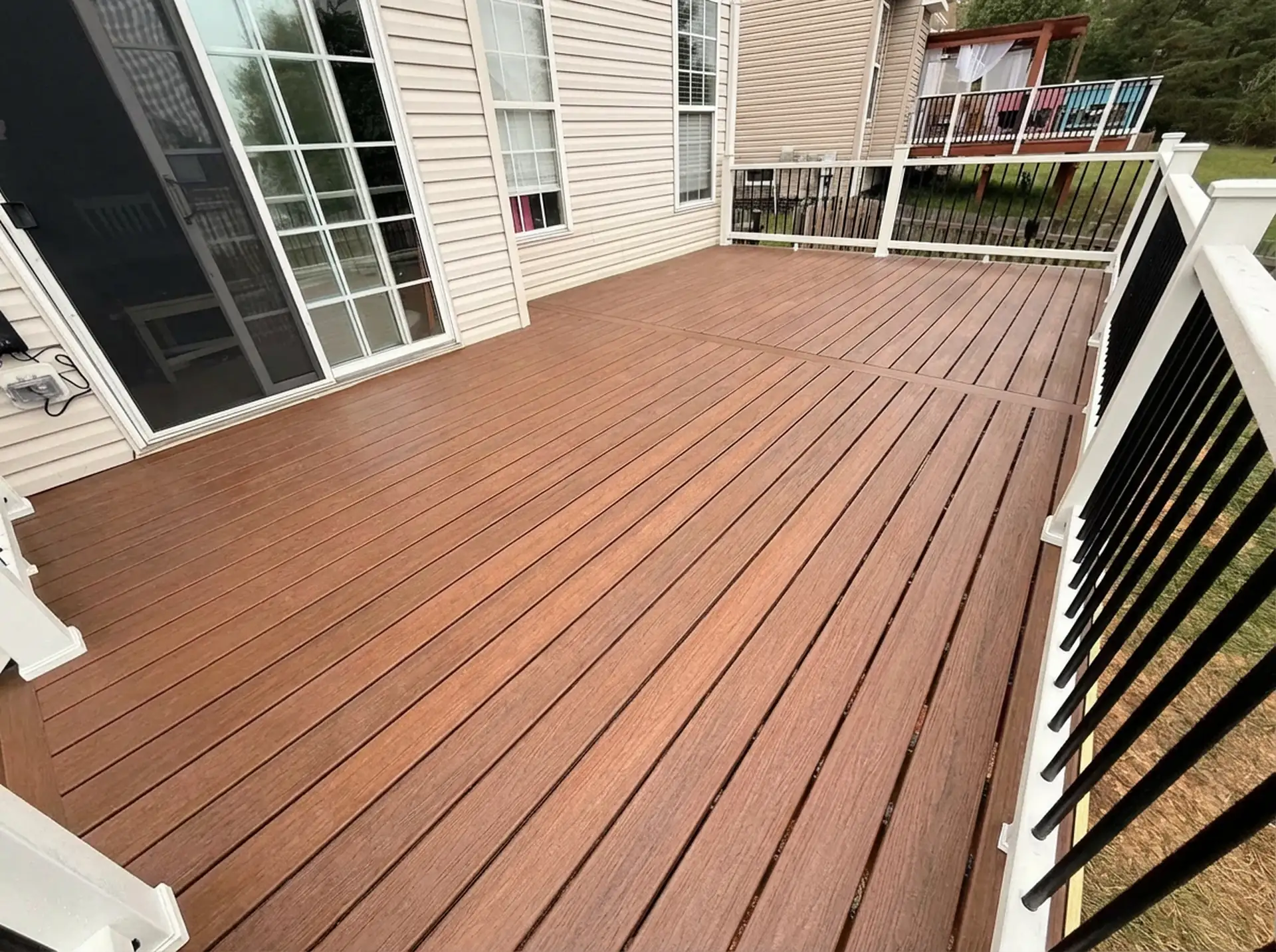 composite decking installation in fredericksburg va A modern outdoor deck made of dark brown composite or treated wood planks. The deck extends from the back of a light-colored, vinyl-sided house. A large sliding glass door opens onto the deck. White railings with black metal balusters line the deck's perimeter.
