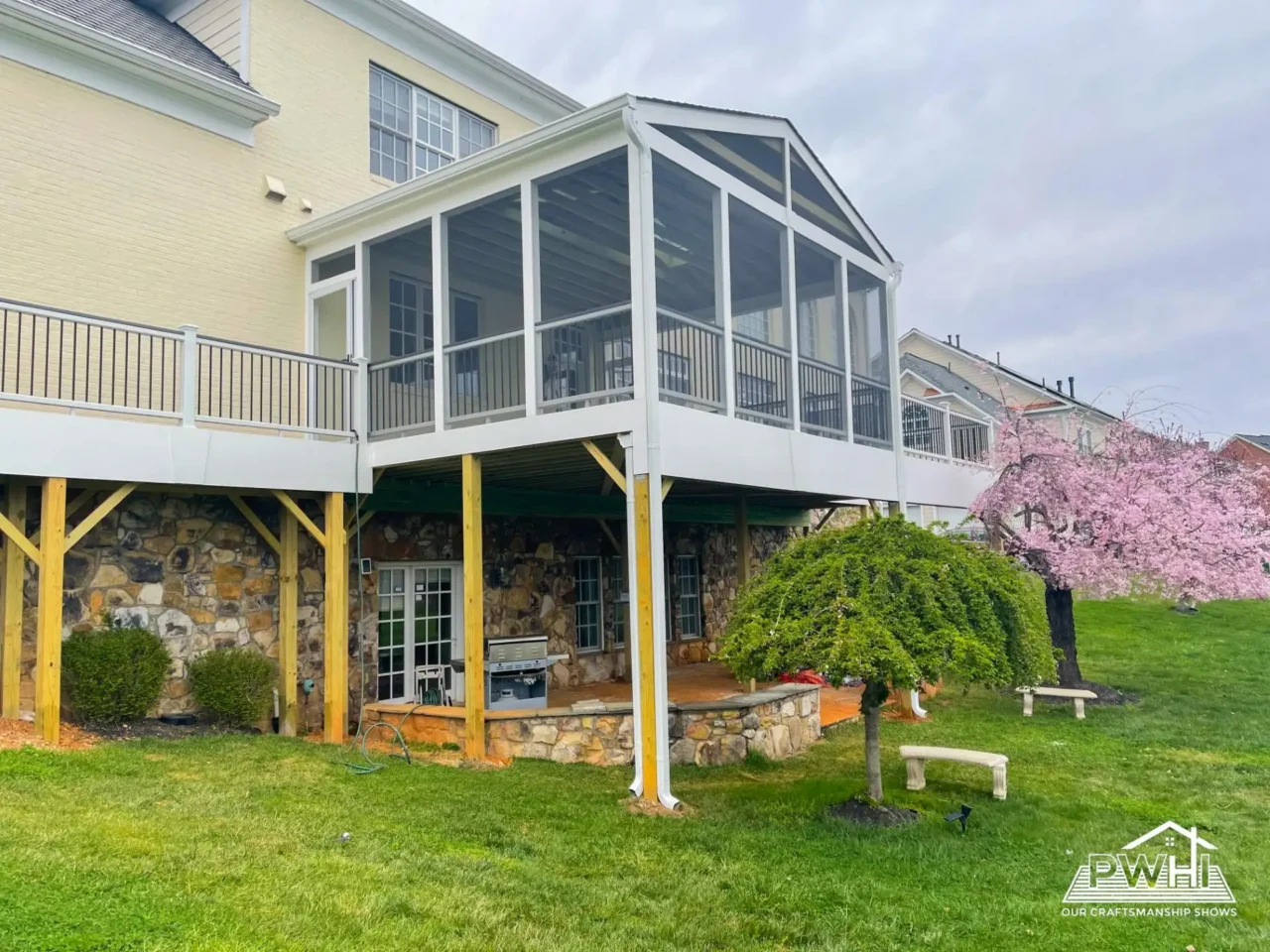 Large white screened-in porch with a pitched roof attached to a yellow house, elevated over a stone patio and supported by wood posts, next to a pink flowering cherry tree and a weeping tree
