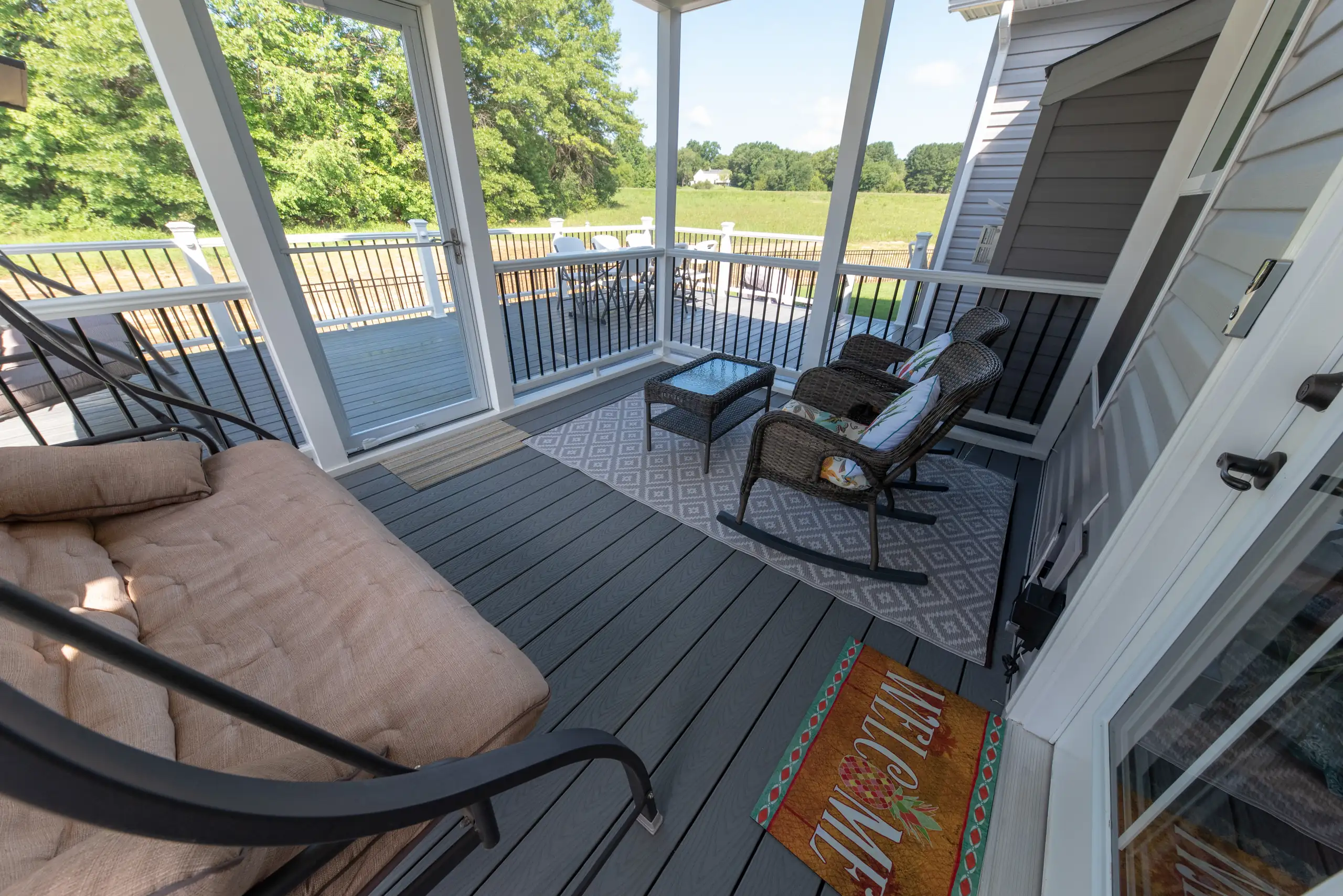 composite deck builder in reston va A screened-in porch with grey composite decking features a large cushioned daybed/swing on the left and two wicker rocking chairs with a glass-top side table on the right, all resting on a patterned outdoor rug. A "WELCOME" doormat is visible near the doorway. The porch overlooks a grassy yard and an open field.