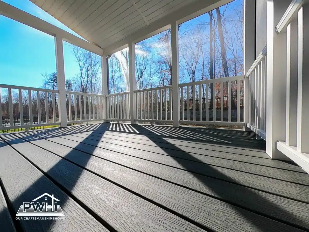 A close-up, low-angle view of a newly constructed covered porch or deck. The decking material is a gray, composite product. The porch features white railings and posts, with a white, sloped roof overhead. Sunlight streams in, casting long, dramatic shadows across the deck boards.