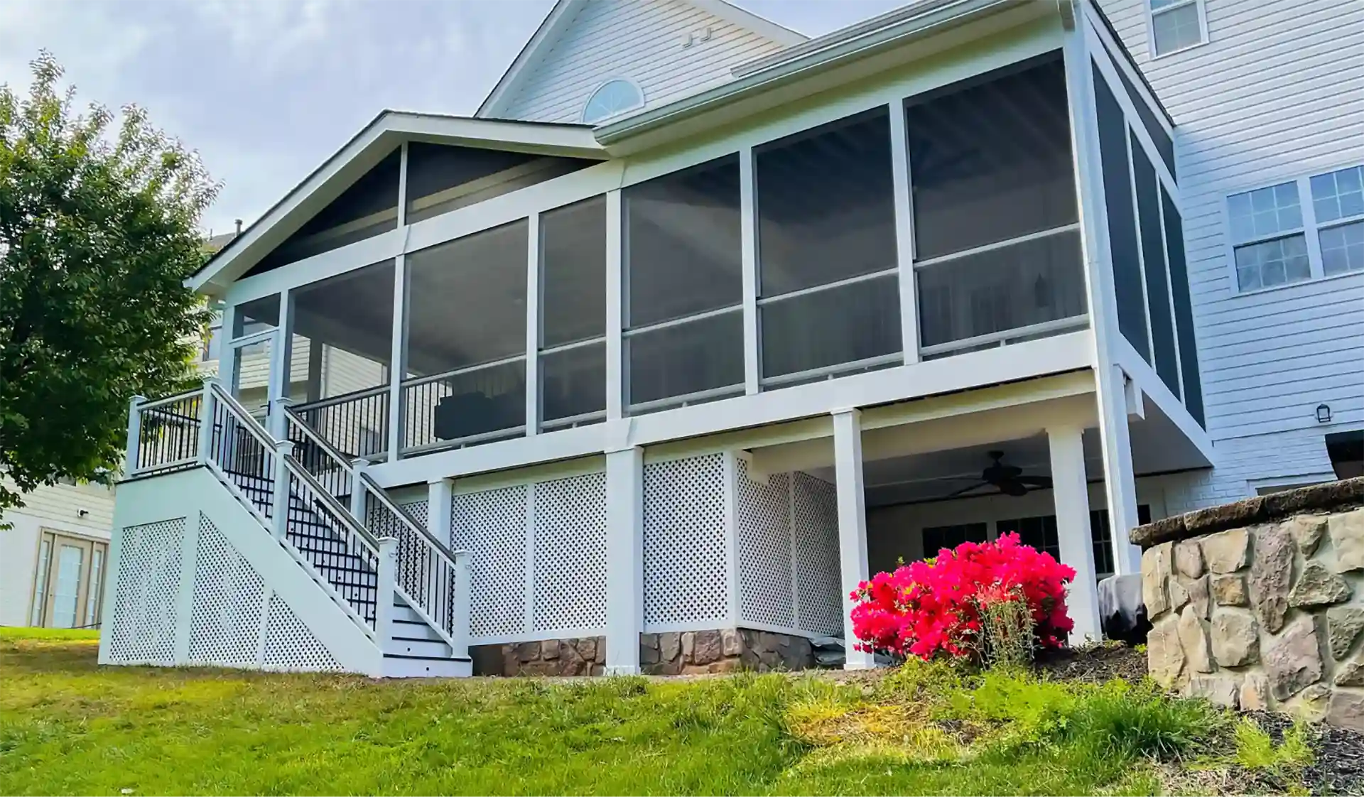 screened porch installation in fairfax va A two-story white house is shown with a large screened-in porch on the back, featuring a set of stairs leading down to the lawn. The lower level of the porch is enclosed by white lattice panels and stone. Bright pink flowers are visible near the porch, and a logo reading "PWH OUR CRAFTSMANSHIP SHOWS" is in the bottom left corner.