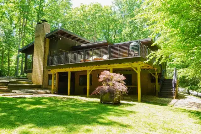 A two-story house with a large wooden deck is nestled in a lush, green, wooded area. The house has a brick chimney and is surrounded by a grassy lawn and tall trees.