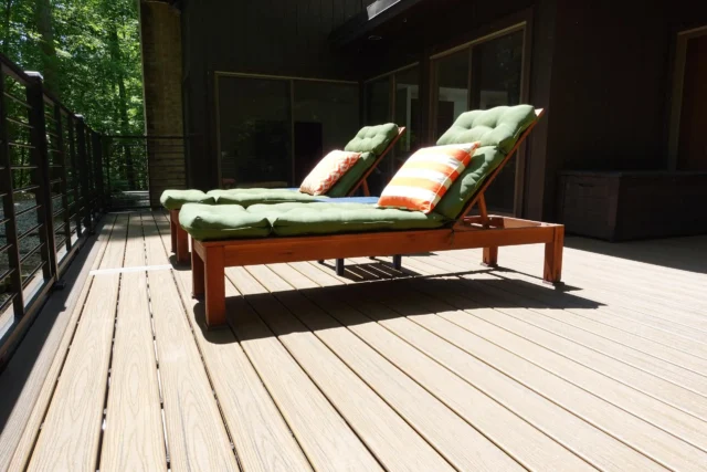 Two wooden lounge chairs with thick, olive-green cushions and striped throw pillows sit on a modern wood-composite deck surrounded by a railing and trees.
