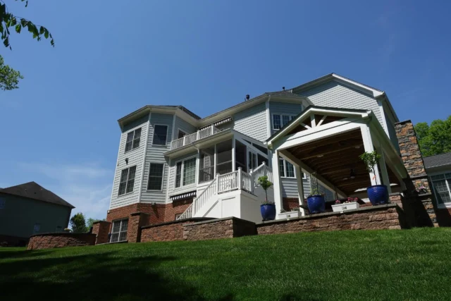 A low-angle exterior view of the back of a large, light-blue house with a brick foundation and accents, featuring a screened-in porch with a white staircase, and a white, covered patio area with dark wood ceiling and large blue planters, all sitting atop a green, sloped lawn.