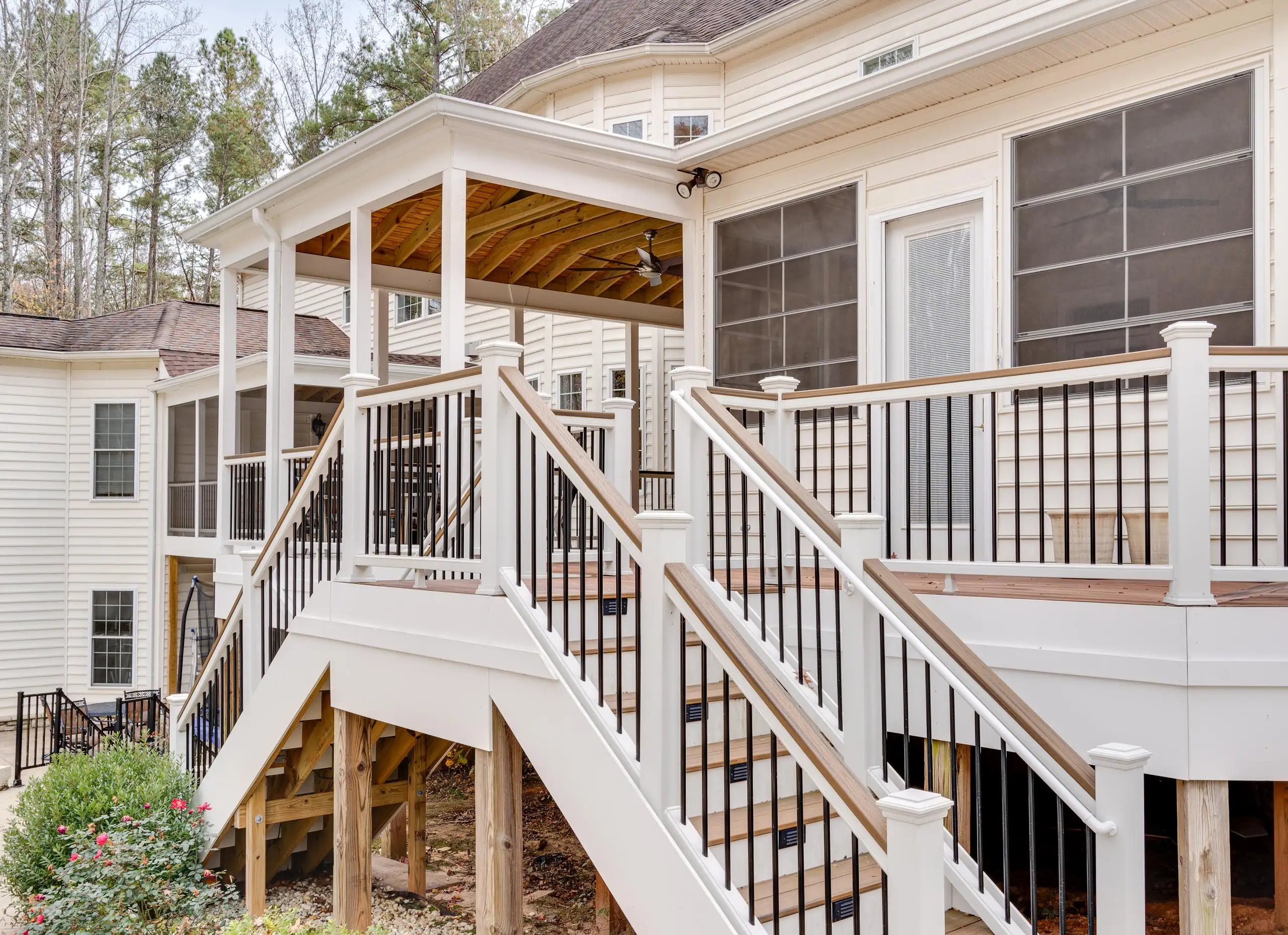 A two-story, cream-colored house with a large deck and white and dark brown railings. The deck has two sets of stairs leading down to the yard. A screened-in porch is visible on the right side of the deck, and a covered porch is on the upper level.