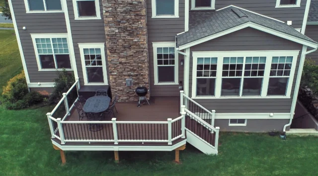 An overhead view of the back of a multi-story, gray house with a stone chimney and a brown deck with white railings. The deck includes an outdoor dining set and a grill.