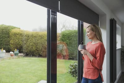 Woman is looking out of black types of patio doors with a cup of tea in her hands.