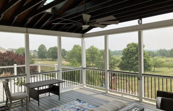 A custom, elevated screened porch addition featuring a dark exposed beam ceiling with skylights, white structural posts, and dark metal railings. The floor is grey composite decking, covered by a blue-and-white patterned rug and furnished with outdoor seating.