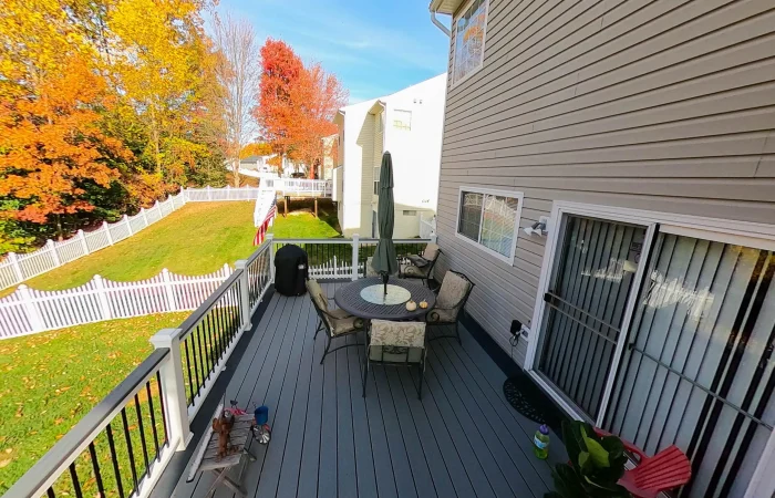 A gray composite deck extends from a tan-sided house on a sunny autumn day. The deck has a white railing with black balusters. Furniture includes a round dining table with an umbrella, four chairs, and a black gas grill. It overlooks a yard with a white picket fence and trees with yellow and red fall foliage.
