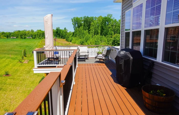 An elevated outdoor deck made of brown composite planks extends from a house with gray siding and large windows. The deck has white railings with black balusters and brown top rails. A black-covered grill stands on the deck near the house. A patio umbrella, a barrel planter, and two lounge chairs are also visible.