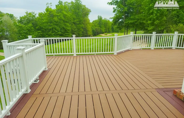 A large composite deck with light brown planks. The deck has a dark reddish-brown border. The deck surface is constructed with two visible sections, with the planks running diagonally toward the center seam from the left and right. The deck is enclosed by a white railing with square posts and vertical balusters.