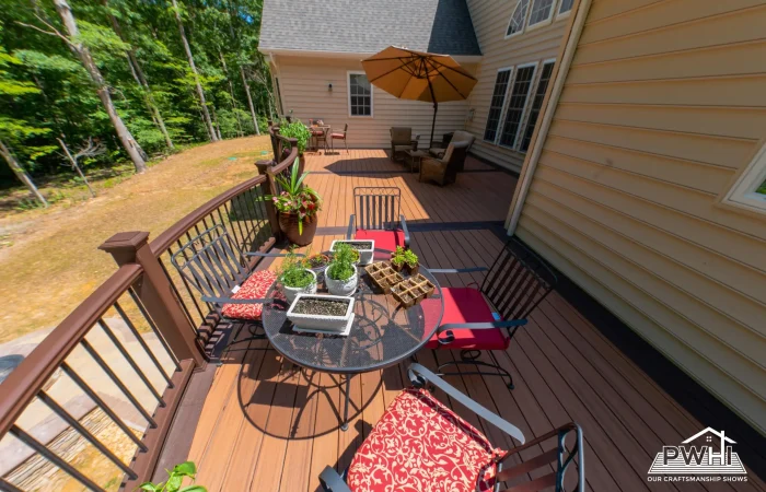 A wide-angle image depicts an outdoor wooden deck with a brown railing, extending from the side of a cream-colored house. The deck is furnished with several pieces of outdoor furniture, including a glass-top table surrounded by chairs with red cushions, and a larger seating area further back with an open umbrella.
